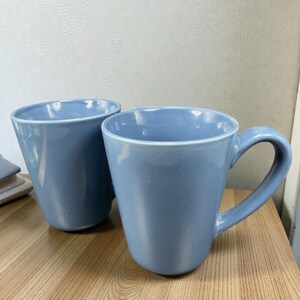 May include: Two light blue ceramic mugs with curved handles sit on a wooden surface. One mug is without a handle. The mugs have a smooth, glossy finish and a simple, modern design. The mugs are likely for coffee or tea.