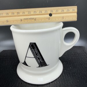 May include: White ceramic mug with a black letter "A" on the front. The mug has a handle and a wide base. A wooden ruler is placed across the top of the mug. The mug is approximately 4 inches tall.