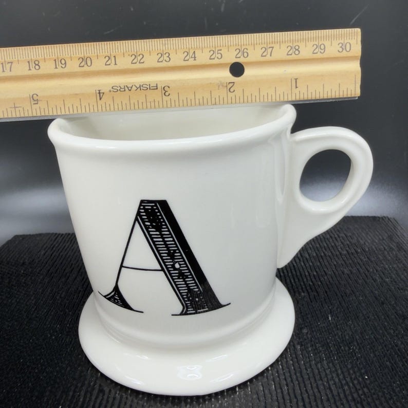 May include: White ceramic mug with a black letter "A" on the front. The mug has a handle and a wide base. A wooden ruler is placed across the top of the mug. The mug is approximately 4 inches tall.