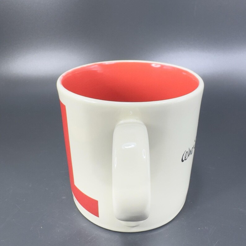 May include: Cream-colored ceramic mug with a red interior and a white handle. A large red "L" is on the side, and the word "Welcome" is written in black script. The mug is designed for hot beverages.