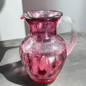 May include: A pink glass pitcher with a clear handle. The pitcher has a textured surface and a rounded body.