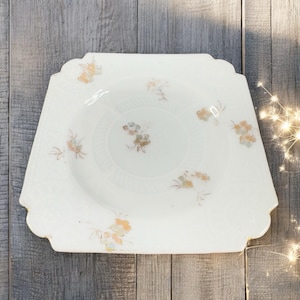 May include: A white, square-shaped plate with a scalloped edge. The plate features a central circular design and is decorated with small floral patterns in shades of orange, yellow, and brown. The plate is set against a wooden background.