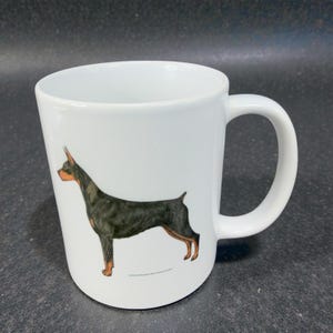 May include: White ceramic mug featuring a Doberman Pinscher illustration. The dog is depicted in black, brown, and tan colors. The mug has a curved shape and a large handle.