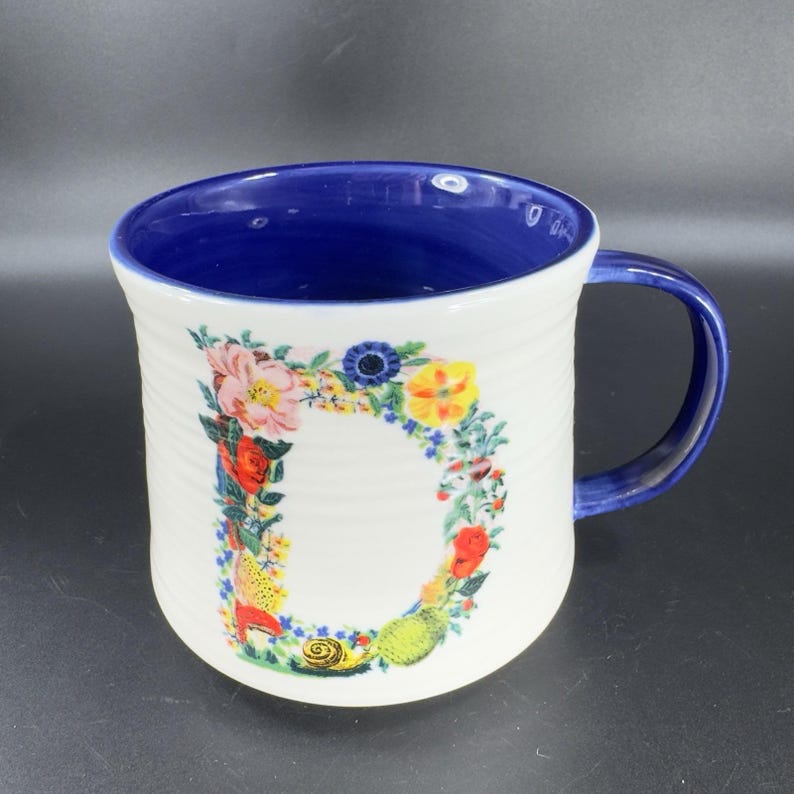 Puede incluir: Taza de cer&aacute;mica blanca con interior y asa azules. La taza presenta un dise&ntilde;o floral en forma de la letra "D" en varios colores. La taza tiene una textura acanalada.