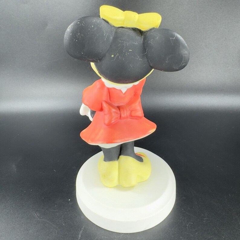 May include: A ceramic figurine of Minnie Mouse standing on a white base. She is wearing a red dress with a yellow bow, black shoes, and a yellow bow in her hair.