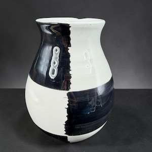 May include: A ceramic vase with a black and white design. The vase has a rounded body and a flared rim. The black paint is applied in a bold, asymmetrical pattern, contrasting with the white background. The vase is suitable for displaying flowers or as a decorative piece.