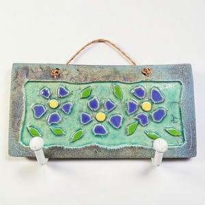 May include: Rectangular ceramic wall hook with a teal and blue floral design. The center features three blue flowers with yellow centers and green leaves. Two white hooks are attached at the bottom, and a woven cord is at the top for hanging.