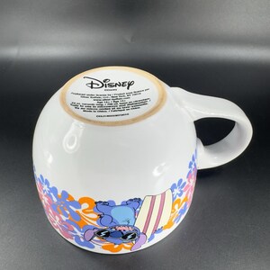 May include: A white ceramic mug with a blue and orange floral design. The mug features a cartoon image of Stitch from Lilo & Stitch, wearing sunglasses and holding a surfboard. The mug has the text "Disney" and "Produced under license by/ Produit sous licence par Silver Buffalo, LLC, New York, NY 10010 www.silverbuffalo.com Age 14+/ Age 14+ Safe to use in microwave and dishwasher.  Va au micro-ondes et au lave-vaisselle. Made in China/ Fabriqu&eacute; en Chine CCL11400338773019"