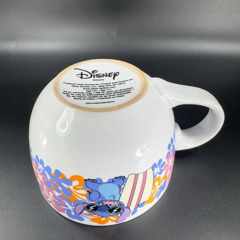 May include: A white ceramic mug with a blue and orange floral design. The mug features a cartoon image of Stitch from Lilo & Stitch, wearing sunglasses and holding a surfboard. The mug has the text "Disney" and "Produced under license by/ Produit sous licence par Silver Buffalo, LLC, New York, NY 10010 www.silverbuffalo.com Age 14+/ Age 14+ Safe to use in microwave and dishwasher.  Va au micro-ondes et au lave-vaisselle. Made in China/ Fabriqu&eacute; en Chine CCL11400338773019"