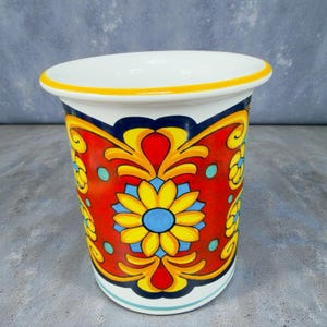 May include: A white ceramic vase with a vibrant floral design. The vase features a red background with yellow and blue floral patterns. The rim is accented with a yellow band. The design is symmetrical and detailed, creating a decorative piece.