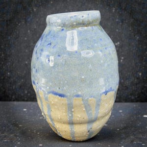 May include: A handmade ceramic vase with a rounded body and a slightly tilted neck. The vase features a light blue glaze with darker blue speckles and vertical drips over a beige base. The pottery has a rustic, textured appearance.