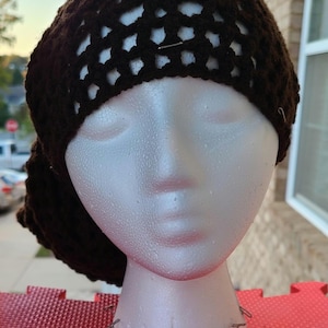 May include: A dark brown crocheted hat with an open weave design on the crown. The hat is displayed on a white mannequin head. The hat's texture and color suggest a handmade item, suitable for various occasions.