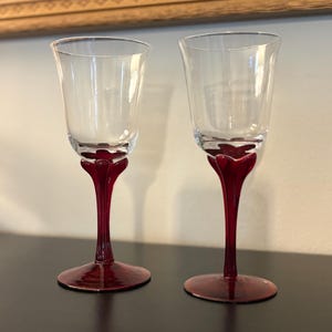 May include: Two clear wine glasses with ruby red stems and bases. The stems have a unique, stylized design. The glasses are displayed on a dark surface, with a painting in the background.