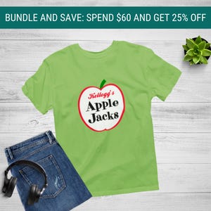 May include: A lime green t-shirt featuring a red-outlined apple graphic with the text Kellogg's Apple Jacks in black, paired with blue denim jeans and black over-ear headphones, with a small green succulent plant nearby.