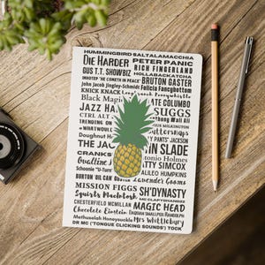 May include: A white notebook featuring a central illustration of a yellow and green pineapple surrounded by a dense collage of black text including names like Die Harder, Peter Panic, and Sh'Dynasty, with a yellow pencil and a silver pen nearby.