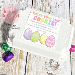 May include: An Easter-themed image showing a white egg carton with a "Paint Your Own Cookie" label. The label has instructions and illustrations of decorated eggs. Chocolate foil-wrapped eggs in various colours surround the carton. The words "Instant Download" are visible.