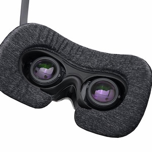 May include: Close-up of a virtual reality headset. The headset features two lenses with a purple reflection, surrounded by a dark gray, textured fabric. The frame is black, and the straps are gray.