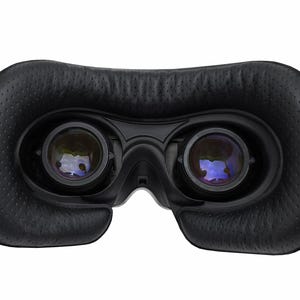 May include: Close-up of a black virtual reality headset. The headset features two lenses, a padded face cushion, and adjustable straps. The lenses are reflective, and the face cushion has a textured surface.