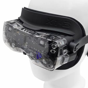 May include: A close-up of a VR headset with a clear, transparent visor revealing internal circuitry. The headset has a black strap and the text "X1-3" is visible. The headset is on a white mannequin head.
