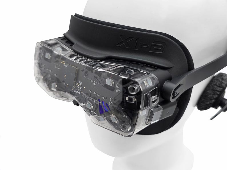 May include: A close-up of a VR headset with a clear, transparent visor revealing internal circuitry. The headset has a black strap and the text "X1-3" is visible. The headset is on a white mannequin head.