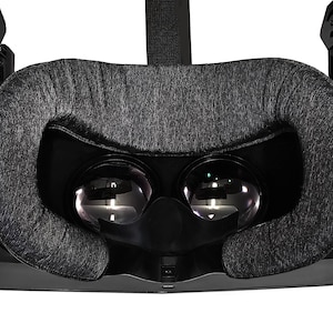 Può includere: Un visore per realtà virtuale nero con un cuscino facciale in tessuto grigio. Il visore ha due lenti e un cinturino nero.