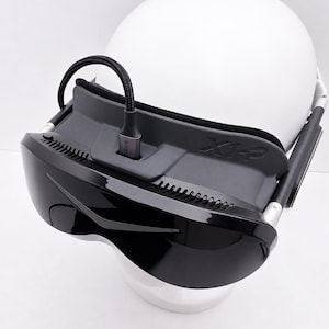 May include: A black virtual reality headset with a glossy front visor and a matte gray frame featuring the text X1-P embossed on the side. A black braided cable is plugged into the top center port of the device, which is mounted on a white head form.