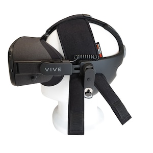 May include: A black VR headset with the word "VIVE" printed on the side. The headset is mounted on a white mannequin head. The headset has adjustable straps and a cushioned headrest for comfort.