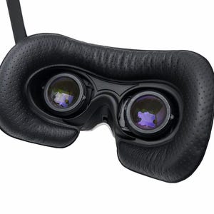 May include: Close-up of a black VR headset with two lenses and a padded face cushion. The lenses reflect a purple hue. The headset has adjustable straps and is designed for virtual reality experiences.