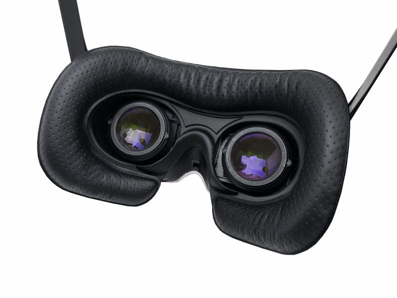 May include: Close-up of a black VR headset with two lenses and a padded face cushion. The lenses reflect a purple hue. The headset has adjustable straps and is designed for virtual reality experiences.
