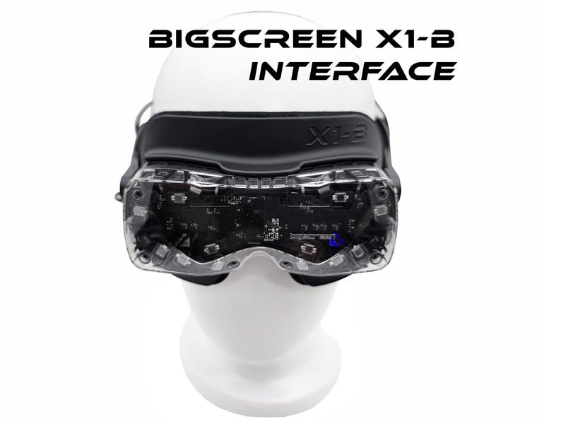 May include: A virtual reality headset with the text "BIGSCREEN X1-B INTERFACE" in black. The headset has a black strap and clear lenses, revealing internal components. It is displayed on a white mannequin head.
