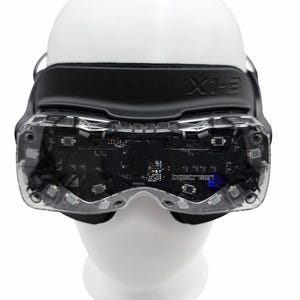 May include: A close-up of a virtual reality headset, model X1-E, with a black strap and clear lenses. The lenses reveal the internal circuitry. The headset is mounted on a white mannequin head.