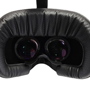 May include: Close-up of a black virtual reality headset with two lenses. The headset has a padded black headband and a black fabric covering the lenses.