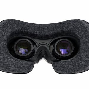 May include: A close-up of a virtual reality headset. The headset features two large lenses, a dark gray fabric face cushion, and black plastic components. The lenses reflect a purple hue. The headset has two black arms extending upwards.