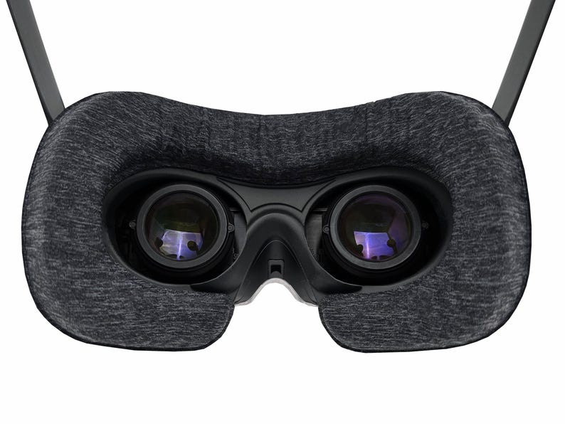 May include: A close-up of a virtual reality headset. The headset features two large lenses, a dark gray fabric face cushion, and black plastic components. The lenses reflect a purple hue. The headset has two black arms extending upwards.