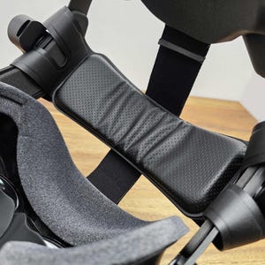 May include: Black VR headset with a padded head strap and a grey fabric lining. The headset has a black adjustable strap and a black padded headrest.