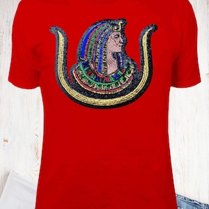May include: Red t-shirt featuring an embroidered design of an Egyptian pharaoh's bust. The pharaoh's head is adorned with a colorful headdress, and the bust is framed by a gold and black decorative element. The shirt is made of a soft material.