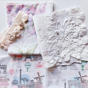 May include: A collection of fabric pieces with floral, lace, and travel patterns. Includes cream lace trim, white fabric with a floral print, and white fabric with a Paris illustration and red bicycles. The fabrics are folded.