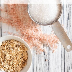 May include: A white bowl filled with rolled oats, a measuring cup filled with Epsom salt, and pink Himalayan salt scattered on a white wooden surface. The text "Oatmeal & Epsom Salt Bath Soak Recipe" is at the top of the image.