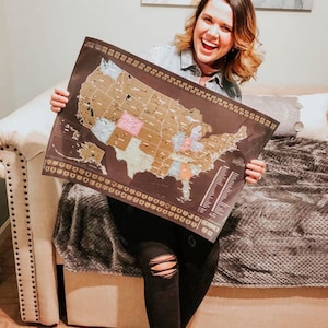 May include: A woman is holding a black and gold scratch-off map of the United States. The map shows all 50 states and is marked with gold foil. The woman is smiling and appears happy.