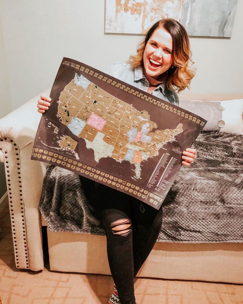 May include: A woman is holding a black and gold scratch-off map of the United States. The map shows all 50 states and is marked with gold foil. The woman is smiling and appears happy.