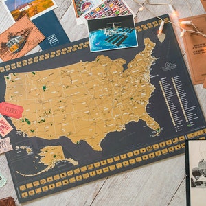 May include: A gold foil scratch-off map of the United States with a black background. The map is labelled "Travelisation" and includes a list of landmarks. The map is surrounded by various travel-themed items, including postcards, a ticket stub, and a string of fairy lights.