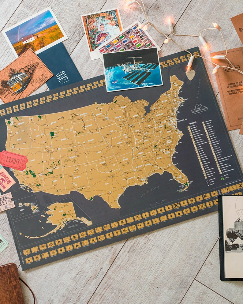 May include: A gold foil scratch-off map of the United States with a black background. The map is labelled "Travelisation" and includes a list of landmarks. The map is surrounded by various travel-themed items, including postcards, a ticket stub, and a string of fairy lights.