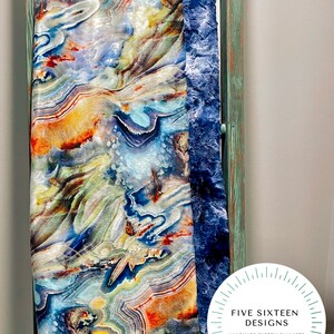 May include: A colorful blanket with an abstract agate design in shades of blue, orange, yellow, and white. The blanket has a plush, dark blue border and is draped over a wooden ladder. The text "FIVE SIXTEEN DESIGNS" is visible.