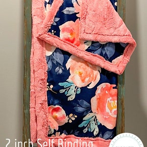 May include: A navy blue floral blanket with peach and white flowers and blue leaves. The blanket has a 2-inch self-binding border in a soft, textured peach material. The blanket is displayed on a wooden ladder.