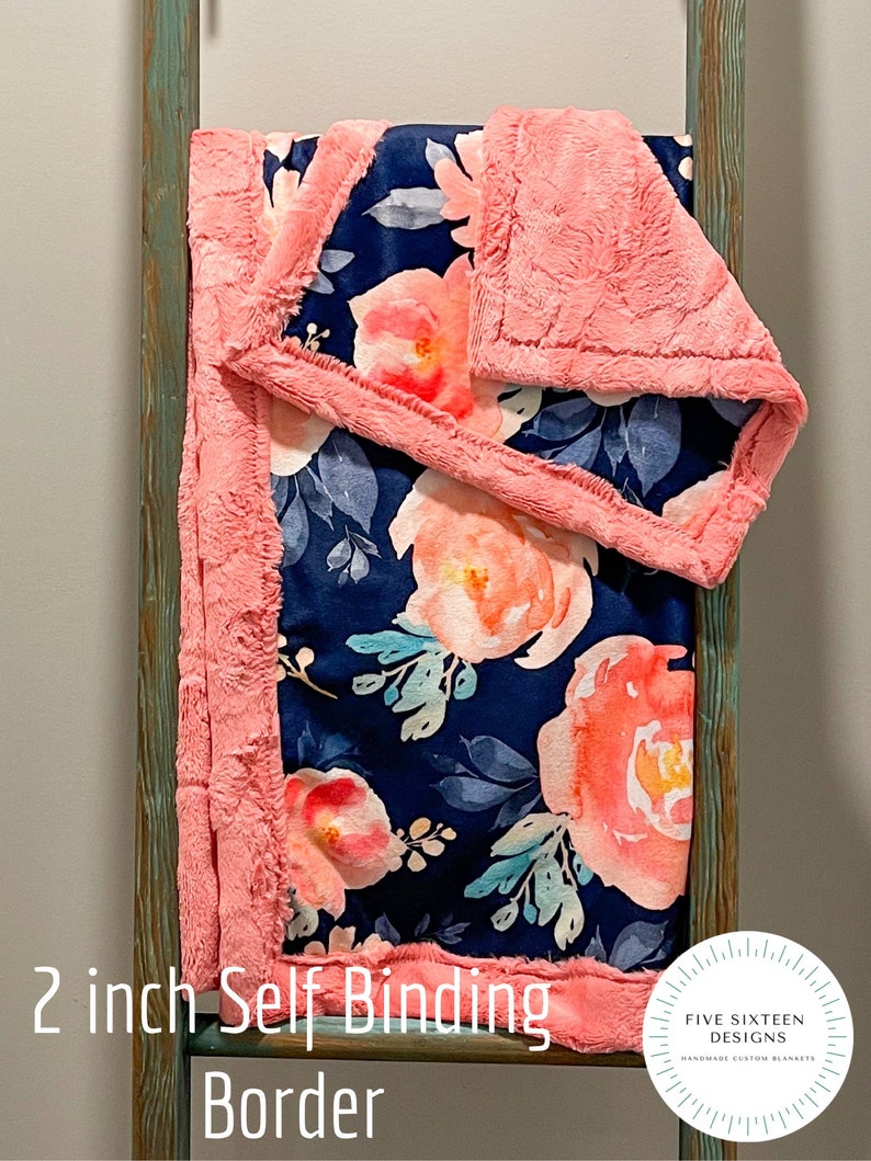 May include: A navy blue floral blanket with peach and white flowers and blue leaves. The blanket has a 2-inch self-binding border in a soft, textured peach material. The blanket is displayed on a wooden ladder.