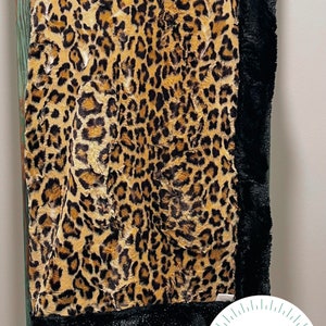 May include: A plush blanket with a leopard print design in shades of tan, brown, and black. The blanket has a black faux fur border. The blanket is draped over a wooden ladder. The blanket is labeled "FIVE SIXTEEN DESIGNS HANDMADE CUSTOM BLANKETS."