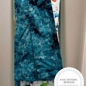 May include: A teal-colored, plush blanket draped over a wooden ladder. The blanket has a textured surface. The bottom edge reveals a patterned fabric with airplanes and a green robot. The blanket is labeled "FIVE SIXTEEN DESIGNS HANDMADE CUSTOM BLANKETS."