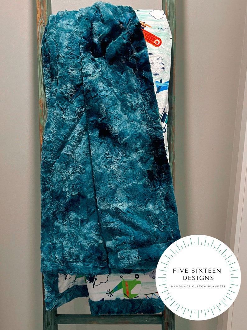 May include: A teal-colored, plush blanket draped over a wooden ladder. The blanket has a textured surface. The bottom edge reveals a patterned fabric with airplanes and a green robot. The blanket is labeled "FIVE SIXTEEN DESIGNS HANDMADE CUSTOM BLANKETS."