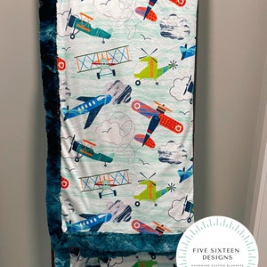 May include: A soft, patterned blanket featuring colorful airplanes, helicopters, and clouds on a light blue background. The blanket has a dark teal plush border and the text "FIVE SIXTEEN DESIGNS HANDMADE CUSTOM BLANKETS" in a circular logo.