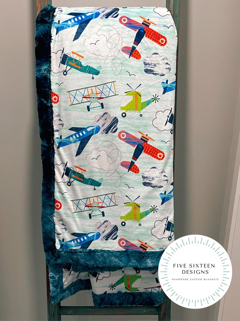 May include: A soft, patterned blanket featuring colorful airplanes, helicopters, and clouds on a light blue background. The blanket has a dark teal plush border and the text "FIVE SIXTEEN DESIGNS HANDMADE CUSTOM BLANKETS" in a circular logo.